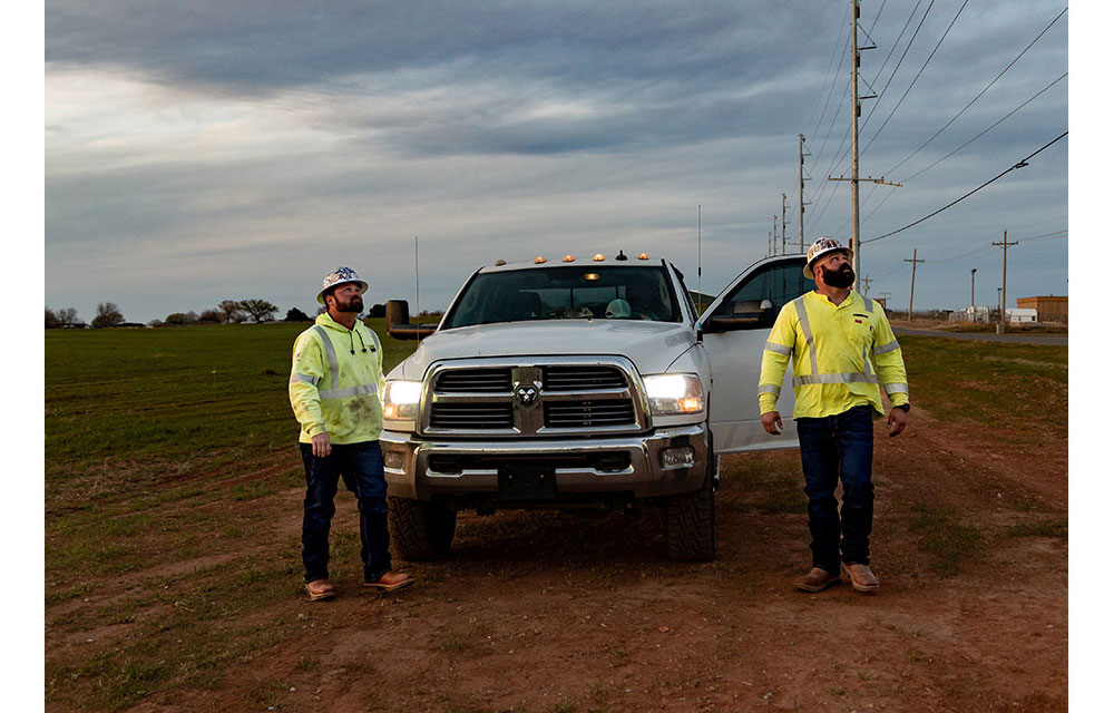 Two electrical linemen in reflective safety gear, Dylan Edgmon and Kevin Walker, stand beside a white work truck with headlights on, parked on a dirt road near power lines.